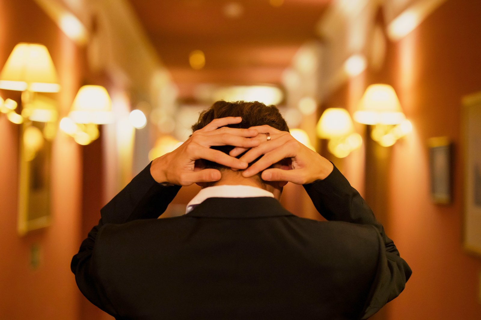 Man in a black suit jacket in a hotel corridor, hands behind his neck after a long evening